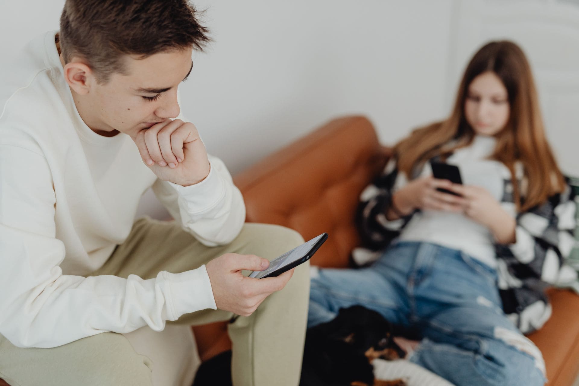 A young man and woman seated apart on a couch, each looking at a smartphone.